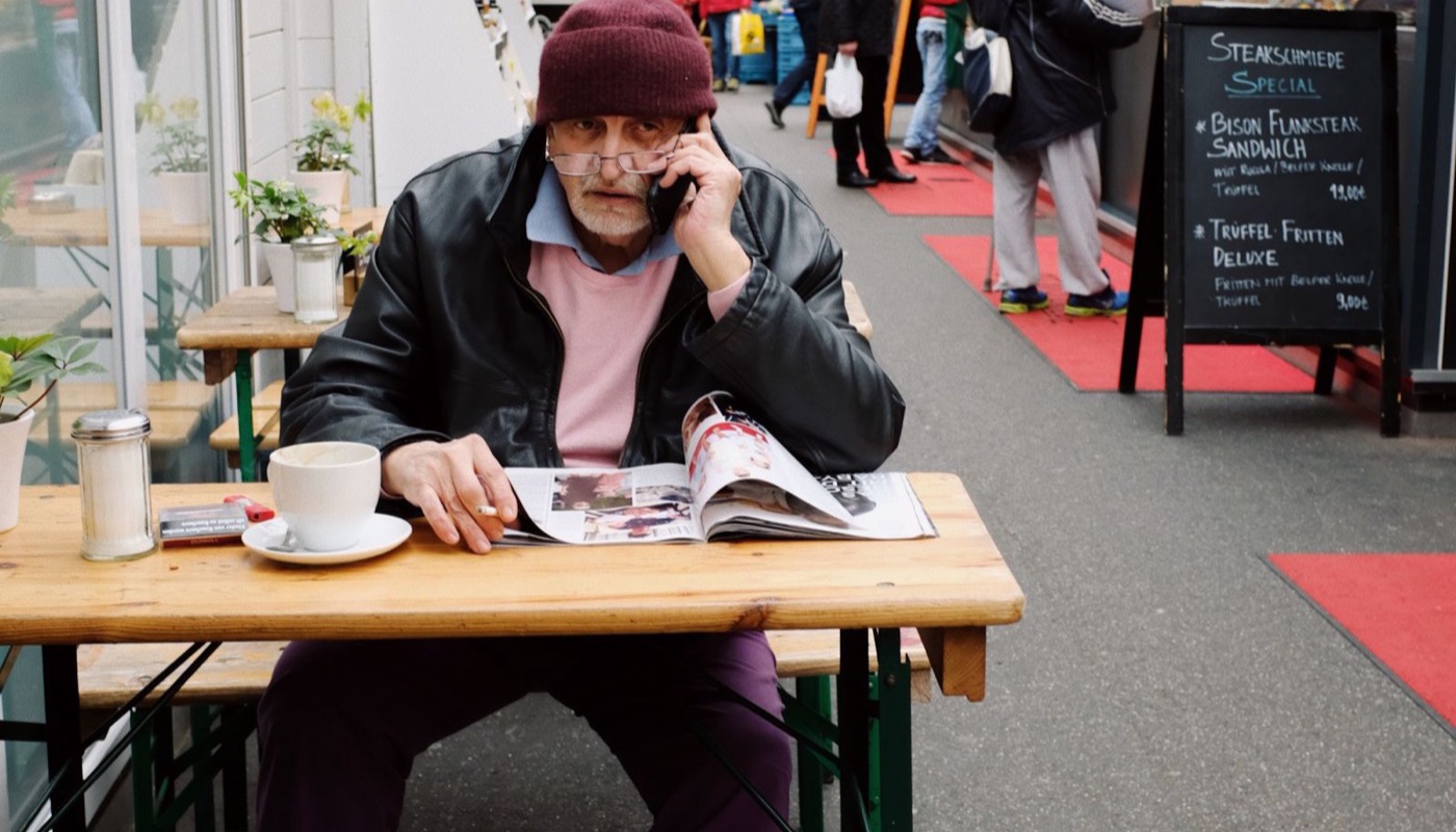 A man at a cafe table reading a newspaper while holding his phone